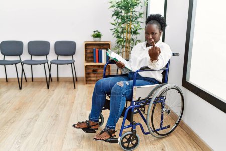 Foto de Young black woman sitting on wheelchair at waiting room showing middle finger, impolite and rude fuck off expression - Imagen libre de derechos