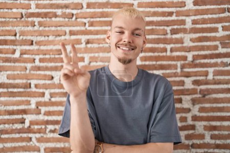 Téléchargez les photos : Jeune homme caucasien debout au-dessus du mur de briques souriant avec le visage heureux clin d'oeil à la caméra faisant signe de victoire avec les doigts. numéro deux. - en image libre de droit