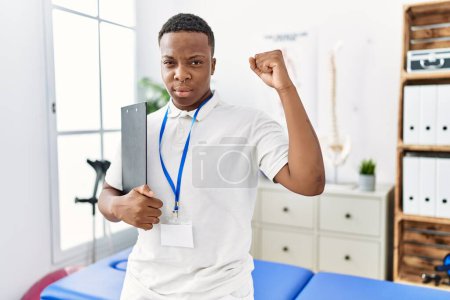 Téléchargez les photos : Jeune homme de physiothérapie africaine travaillant à la clinique de récupération de la douleur agacé et frustré criant de colère, criant fou de colère et la main levée - en image libre de droit