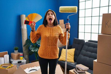 Photo for Young hispanic woman painting home walls with paint roller angry and mad screaming frustrated and furious, shouting with anger looking up. - Royalty Free Image