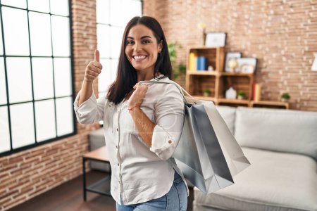 Foto de Mujer hispana joven sosteniendo bolsas de compras sonriendo feliz y positivo, pulgar hacia arriba haciendo excelente y signo de aprobación - Imagen libre de derechos
