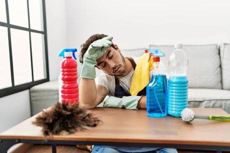 Photo for Young hispanic man tired leaning on table with cleaning products at home - Royalty Free Image