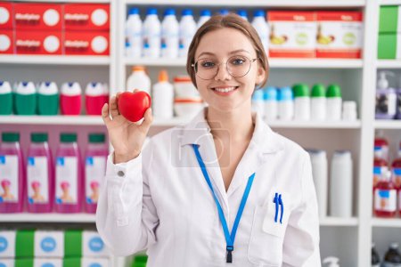 Téléchargez les photos : Jeune femme caucasienne travaillant à la pharmacie pharmacie tenant coeur rouge regardant positif et heureux debout et souriant avec un sourire confiant montrant des dents - en image libre de droit