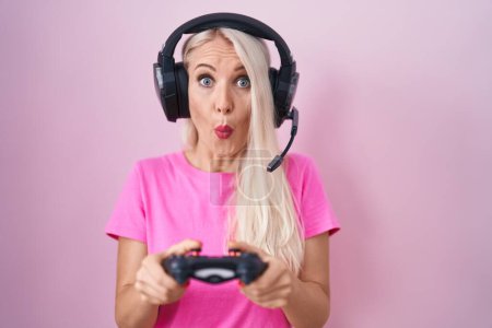Photo for Caucasian woman playing video game holding controller in shock face, looking skeptical and sarcastic, surprised with open mouth - Royalty Free Image