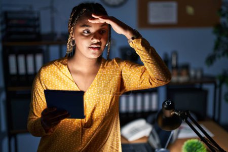 Téléchargez les photos : Femme afro-américaine avec des tresses travaillant au bureau la nuit avec une tablette très heureuse et souriante regardant loin avec la main au-dessus de la tête. concept de recherche. - en image libre de droit