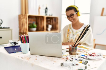 Foto de Mujer hispana joven escuchando música usando un portátil y dibujando en un cuaderno en un estudio de arte - Imagen libre de derechos