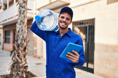 Photo for Young hispanic man courier holding water bottle using touchpad at street - Royalty Free Image