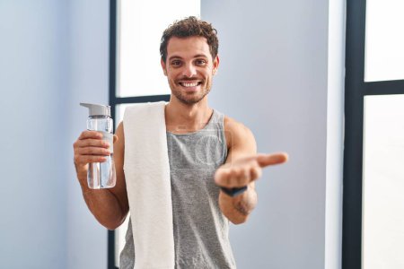 Photo for Young hispanic man wearing sportswear drinking water smiling cheerful offering palm hand giving assistance and acceptance. - Royalty Free Image