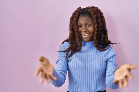 Téléchargez les photos : Femme africaine debout sur fond rose souriant joyeuse offrant des mains donnant assistance et acceptation. - en image libre de droit