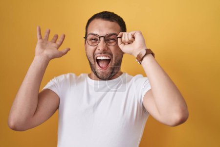 Téléchargez les photos : Homme hispanique tenant des lunettes célébrant la victoire avec un sourire heureux et l'expression gagnante avec les mains levées - en image libre de droit