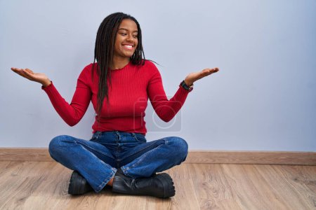 Photo for Young african american with braids sitting on the floor at home smiling showing both hands open palms, presenting and advertising comparison and balance - Royalty Free Image