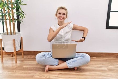 Photo for Young blonde woman using computer laptop sitting on the floor at the living room gesturing with hands showing big and large size sign, measure symbol. smiling looking at the camera. measuring concept. - Royalty Free Image
