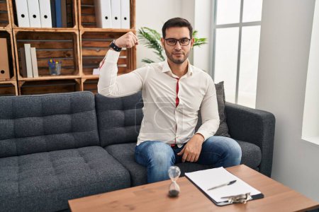 Photo for Young hispanic man with beard working at consultation office strong person showing arm muscle, confident and proud of power - Royalty Free Image