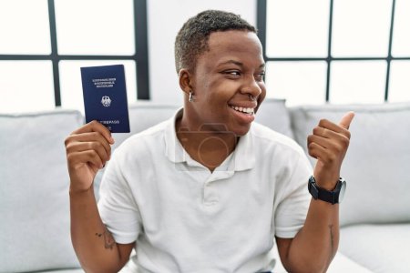 Téléchargez les photos : Jeune homme africain tenant un passeport deutschland pointant le pouce vers le côté souriant heureux avec la bouche ouverte - en image libre de droit