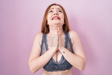 Foto de Redhead woman wearing lingerie over pink background begging and praying with hands together with hope expression on face very emotional and worried. begging. - Imagen libre de derechos