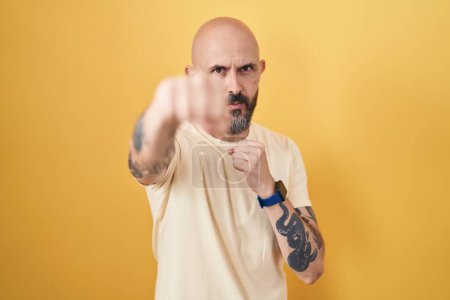 Photo for Hispanic man with tattoos standing over yellow background punching fist to fight, aggressive and angry attack, threat and violence - Royalty Free Image