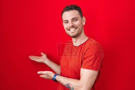 Téléchargez les photos : Jeune homme hispanique debout sur fond rouge invitant à entrer souriant naturel avec la main ouverte - en image libre de droit