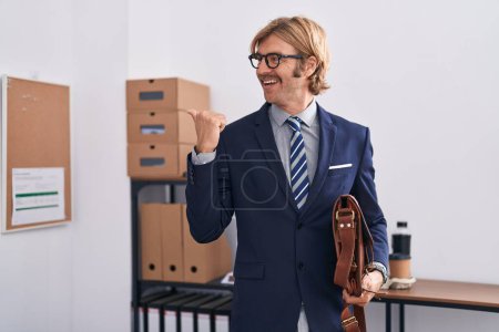 Photo for Caucasian man with mustache working at the office wearing glasses pointing thumb up to the side smiling happy with open mouth - Royalty Free Image