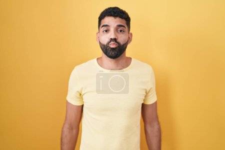 Téléchargez les photos : Homme hispanique avec barbe debout sur fond jaune regardant la caméra souffler un baiser sur l'air étant belle et sexy. expression d'amour. - en image libre de droit