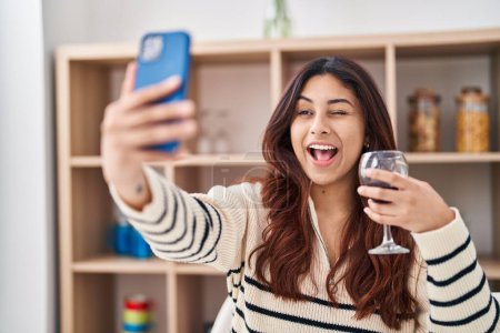 Photo for Hispanic young business woman taking a selfie picture drinking a glass of wine winking looking at the camera with sexy expression, cheerful and happy face. - Royalty Free Image