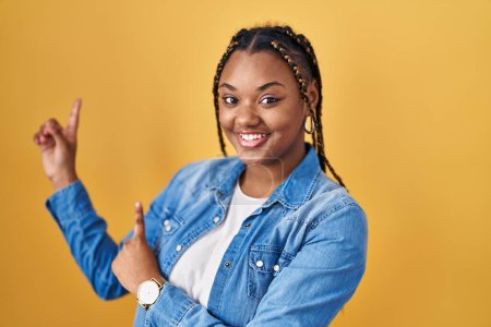 Photo for African american woman with braids standing over yellow background smiling and looking at the camera pointing with two hands and fingers to the side. - Royalty Free Image