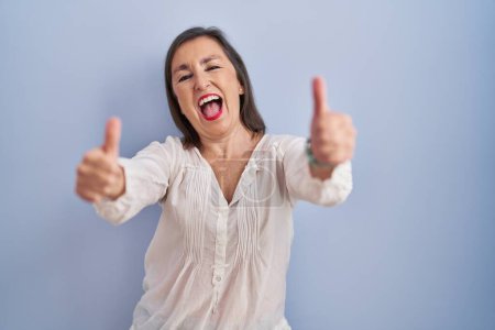 Foto de Mujer hispana de mediana edad de pie sobre fondo azul aprobando hacer gesto positivo con la mano, pulgares hacia arriba sonriendo y feliz por el éxito. gesto ganador. - Imagen libre de derechos