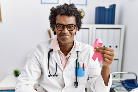Foto de African doctor man holding pink cancer ribbon looking positive and happy standing and smiling with a confident smile showing teeth - Imagen libre de derechos
