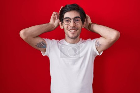 Foto de Joven hombre hispano de pie sobre fondo rojo posando divertido y loco con los dedos en la cabeza como orejas de conejo, sonriendo alegre - Imagen libre de derechos
