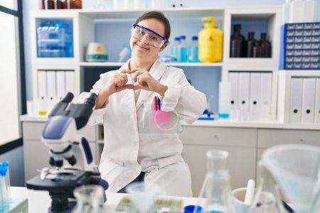 Foto de Chica hispana con síndrome de Down que trabaja en el laboratorio científico sonriendo en el amor haciendo forma de símbolo del corazón con las manos. concepto romántico. - Imagen libre de derechos