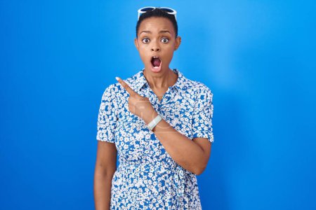Foto de African american woman standing over blue background surprised pointing with finger to the side, open mouth amazed expression. - Imagen libre de derechos