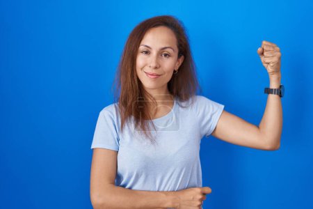Téléchargez les photos : Brunette woman standing over blue background strong person showing arm muscle, confident and proud of power - en image libre de droit