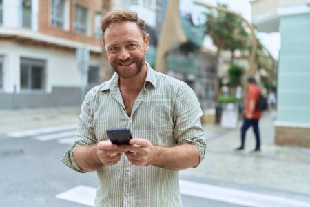 Téléchargez les photos : Moyen Age homme souriant confiant en utilisant smartphone à la rue - en image libre de droit