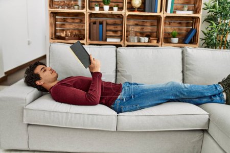 Foto de Joven hispano leyendo libro tumbado en el sofá de su casa. - Imagen libre de derechos