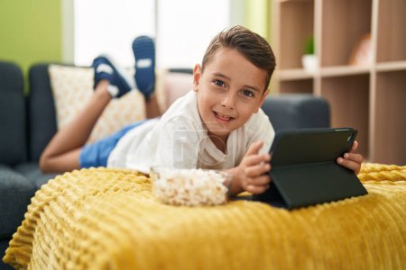 Téléchargez les photos : Adorable bambin hispanique regardant la vidéo sur le pavé tactile couché sur le canapé à la maison - en image libre de droit