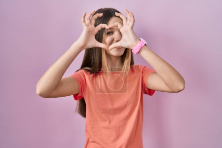Foto de Teenager girl standing over pink background doing heart shape with hand and fingers smiling looking through sign - Imagen libre de derechos