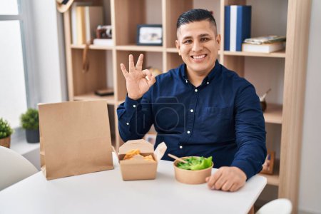Foto de Hispano joven comiendo llevar comida haciendo ok signo con los dedos, sonriendo gesto amistoso excelente símbolo - Imagen libre de derechos