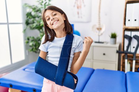 Photo for Young hispanic girl wearing arm on sling at rehabilitation clinic screaming proud, celebrating victory and success very excited with raised arms - Royalty Free Image