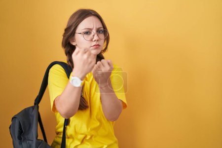 Photo for Young caucasian woman wearing student backpack over yellow background ready to fight with fist defense gesture, angry and upset face, afraid of problem - Royalty Free Image