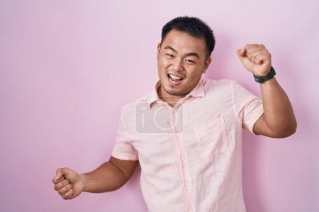 Téléchargez les photos : Jeune homme chinois debout sur fond rose dansant heureux et joyeux, souriant décontracté et confiant écoutant de la musique - en image libre de droit