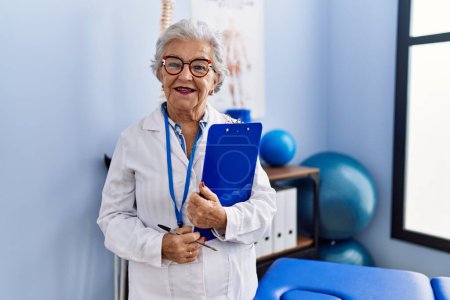 Foto de Mujer mayor de cabello gris que usa uniforme de fisioterapeuta con lista de verificación en la clínica de fisioterapia - Imagen libre de derechos