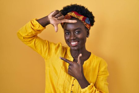 Téléchargez les photos : Jeune femme africaine portant turban africain souriant faisant cadre avec les mains et les doigts avec le visage heureux. concept de créativité et photographie. - en image libre de droit