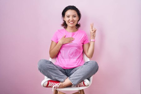 Téléchargez les photos : Jeune femme hispanique assise sur une chaise sur un fond rose en jurant avec la main sur la poitrine et les doigts vers le haut, faisant un serment de fidélité - en image libre de droit