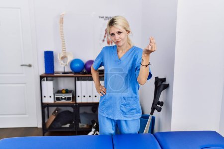 Foto de Joven fisioterapeuta mujer que trabaja en la clínica de recuperación del dolor haciendo un gesto italiano con la mano y los dedos expresión de confianza - Imagen libre de derechos