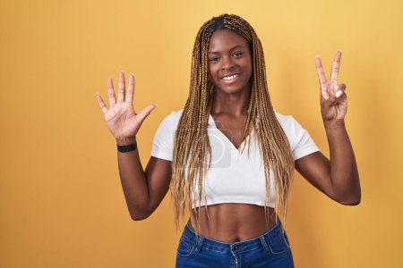 Foto de Mujer afroamericana con el pelo trenzado de pie sobre fondo amarillo mostrando y apuntando hacia arriba con los dedos número siete mientras sonríe confiado y feliz. - Imagen libre de derechos