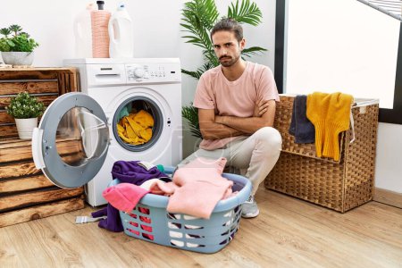 Foto de Young hispanic man putting dirty laundry into washing machine skeptic and nervous, disapproving expression on face with crossed arms. negative person. - Imagen libre de derechos