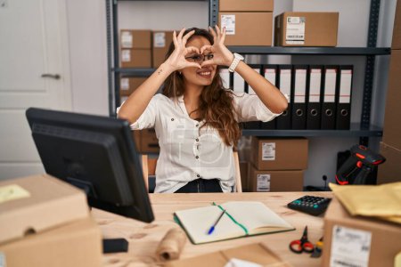 Téléchargez les photos : Young hispanic woman working at small business ecommerce doing heart shape with hand and fingers smiling looking through sign - en image libre de droit