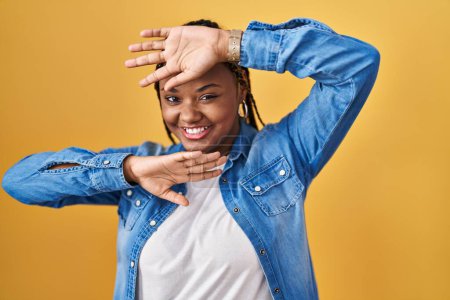Photo for African american woman with braids standing over yellow background smiling cheerful playing peek a boo with hands showing face. surprised and exited - Royalty Free Image
