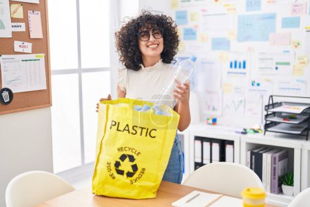 Photo for Young middle east woman holding recycling bag with plastic bottles at the office winking looking at the camera with sexy expression, cheerful and happy face. - Royalty Free Image