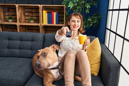 Photo for Young caucasian woman drinking coffee and watching movie sitting on sofa with dog at home - Royalty Free Image