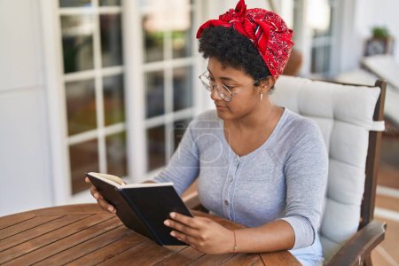 Téléchargez les photos : Femme afro-américaine livre de lecture assis sur la table à la maison terrasse - en image libre de droit